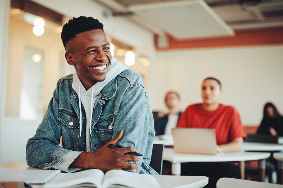 Specialized Business Insurance - Smiling Male Student Sitting in University Classroom with Classmates Blurred at Their Desks in the Background